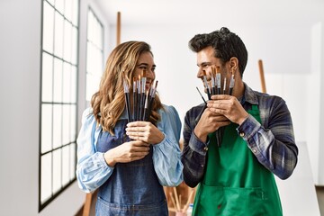 Two hispanic students smiling happy covering face with paintbrushes at art school.