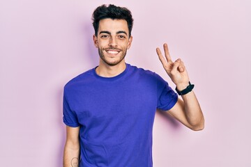 Young hispanic man wearing casual t shirt smiling looking to the camera showing fingers doing victory sign. number two.