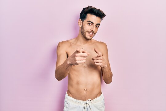 Young hispanic man wearing swimwear shirtless pointing fingers to camera with happy and funny face. good energy and vibes.
