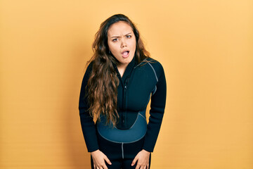 Young hispanic girl wearing diver neoprene uniform in shock face, looking skeptical and sarcastic,...