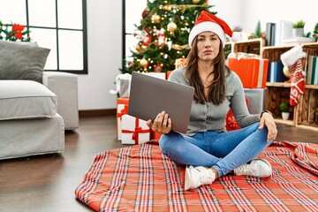 Young latin woman using laptop sitting by christmas tree skeptic and nervous, frowning upset because of problem. negative person.