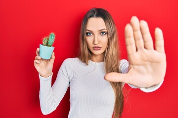 Young blonde girl holding small cactus pot with open hand doing stop sign with serious and...