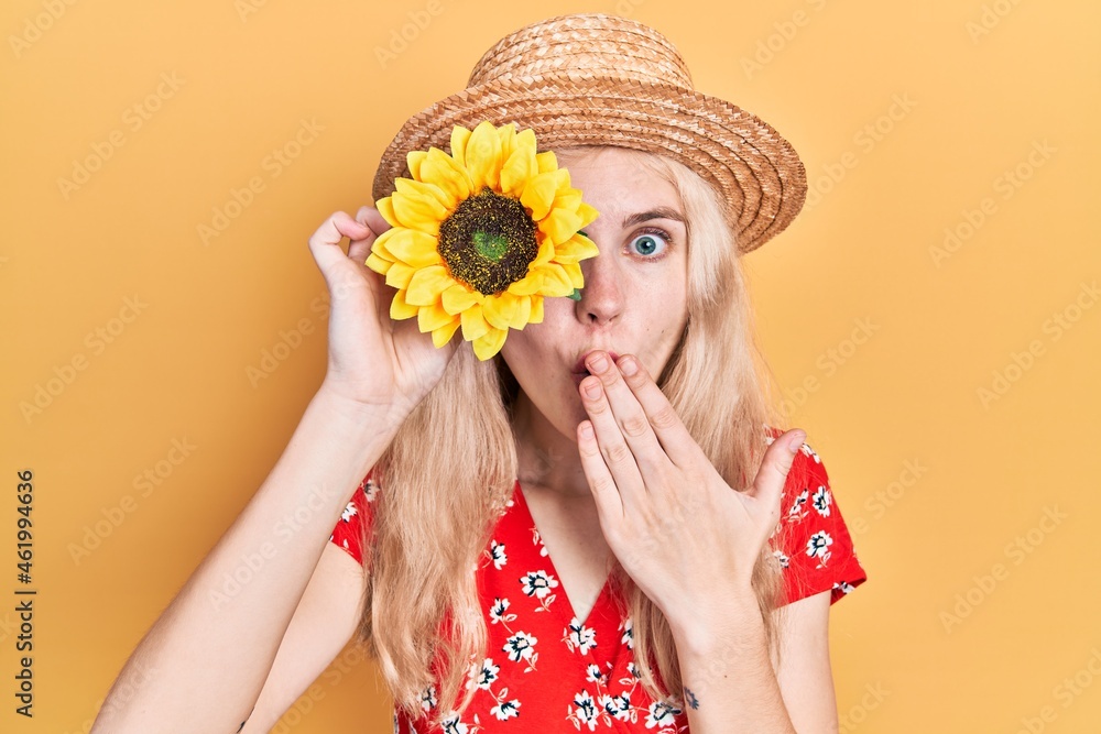 Sticker Beautiful caucasian woman with blond hair holding yellow sunflowers over eye covering mouth with hand, shocked and afraid for mistake. surprised expression