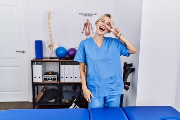 Young physiotherapist woman working at pain recovery clinic doing peace symbol with fingers over face, smiling cheerful showing victory