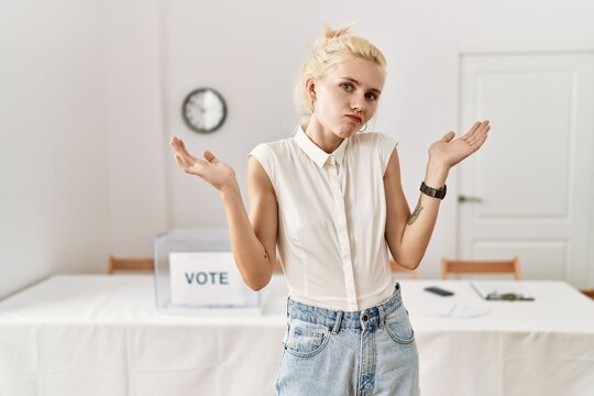 Beautiful Caucasian Woman Standing By Voting Ballot At Election Room Clueless And Confused Expression With Arms And Hands Raised. Doubt Concept.