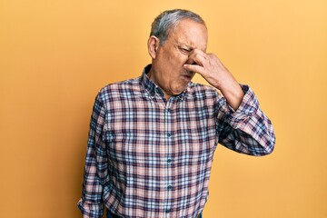 Handsome senior man with grey hair wearing casual shirt smelling something stinky and disgusting, intolerable smell, holding breath with fingers on nose. bad smell