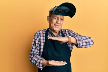 Handsome senior man with grey hair wearing welding protection mask gesturing with hands showing big and large size sign, measure symbol. smiling looking at the camera. measuring concept.