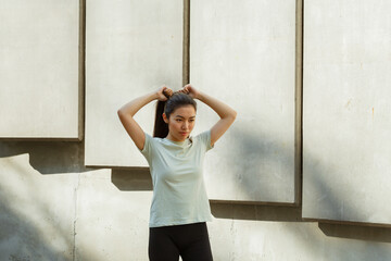 Fototapeta premium Pretty Asian young woman in t-shirt adjusts ponytail made of long dark hair standing near white wall on sunny day middle view