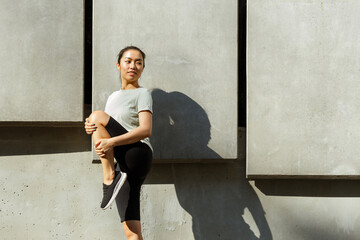 Young Asian sportswoman in tracksuit and sneakers does exercises for legs bending knee at training near wall casting shadow on street