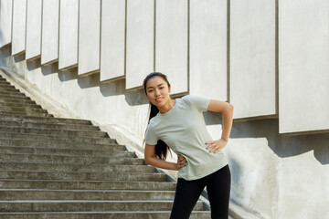 Slim young Asian woman in tracksuit holds hands on waist at training near empty stone steps and concrete wall in city street