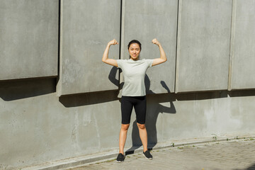 Fototapeta na wymiar Happy brunette Asian sportswoman in light t-shirt shows arm muscles standing by wall with concrete plates on street