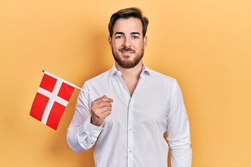 Handsome caucasian man with beard holding denmark flag looking positive and happy standing and...