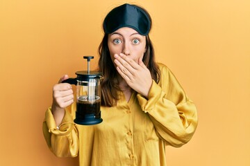 Young brunette woman wearing sleep mask holding coffee maker covering mouth with hand, shocked and...
