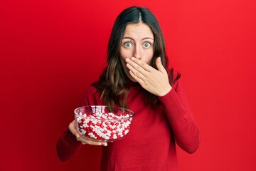 Young brunette woman holding bowl full of pills covering mouth with hand, shocked and afraid for mistake. surprised expression