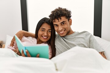 Young latin couple reading book sitting on the bed at bedroom.