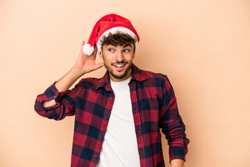 Young arab man celebrating Christmas isolated on beige background trying to listening a gossip.