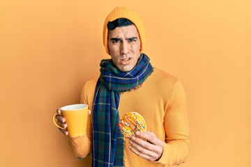 Handsome hispanic man eating doughnut and drinking coffee clueless and confused expression. doubt concept.