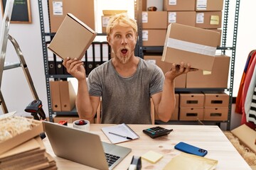Young blond man holding packages working at online shop afraid and shocked with surprise and amazed...