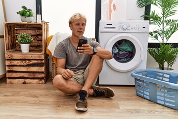 Young blond man doing laundry using smartphone winking looking at the camera with sexy expression,...