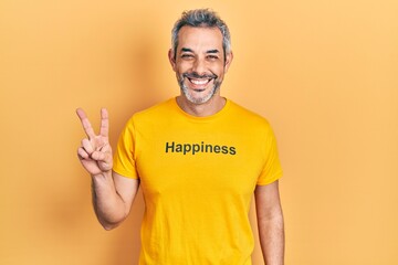 Handsome middle age man with grey hair wearing t shirt with happiness word message smiling with happy face winking at the camera doing victory sign. number two.
