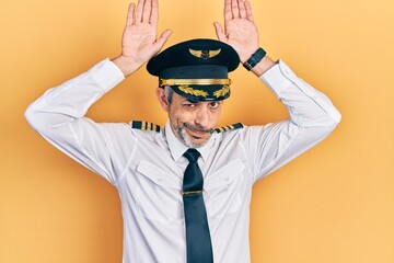 Handsome middle age man with grey hair wearing airplane pilot uniform doing bunny ears gesture with...