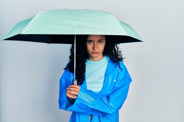Young hispanic woman with curly hair wearing a raincoat and umbrella skeptic and nervous, frowning...