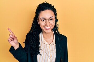 Young hispanic woman with curly hair wearing call center agent headset smiling happy pointing with hand and finger to the side