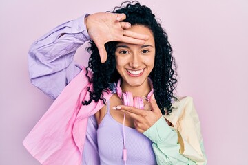 Young hispanic woman with curly hair wearing gym clothes and using headphones smiling making frame with hands and fingers with happy face. creativity and photography concept.