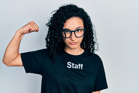Young hispanic woman with curly hair wearing staff t shirt strong person showing arm muscle, confident and proud of power