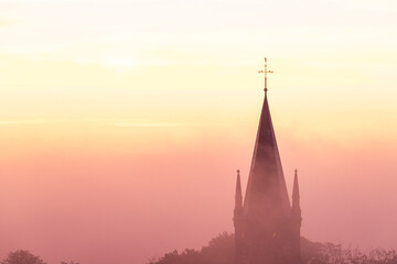 Fototapeta na wymiar A typical colorful Autumn sunrise in Maastricht with the landscape covered with a layer of fog, leaving only silhouettes visible in the distance, like this tower of a church on the hillside.