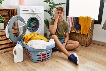 Young caucasian man putting dirty laundry into washing machine covering one eye with hand, confident smile on face and surprise emotion.