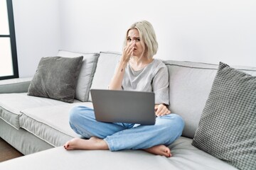 Young caucasian woman using laptop at home sitting on the sofa smelling something stinky and disgusting, intolerable smell, holding breath with fingers on nose. bad smell - obrazy, fototapety, plakaty
