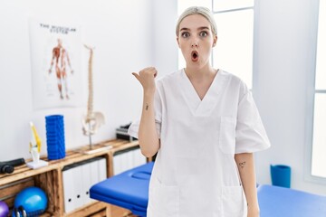 Young caucasian woman working at pain recovery clinic surprised pointing with hand finger to the side, open mouth amazed expression.