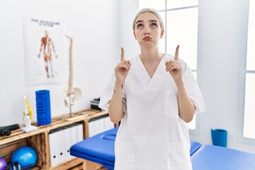 Fototapeta premium Young caucasian woman working at pain recovery clinic pointing up looking sad and upset, indicating direction with fingers, unhappy and depressed.