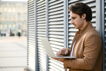 Side view of businessman in coat using laptop near building on street 