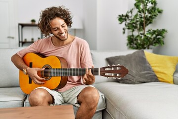 Young hispanic man smiling happy playing classical guitar sitting on the sofa at home.