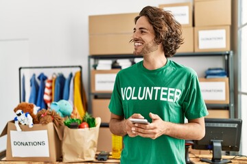 Young hispanic volunteer man smiling happy using smartphone at charity center.