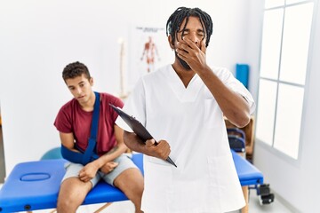 Young hispanic man working at pain recovery clinic with a man with broken arm bored yawning tired covering mouth with hand. restless and sleepiness.