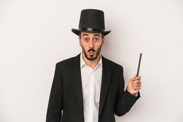 Young magician man holding wand isolated on white background shrugs shoulders and open eyes confused.