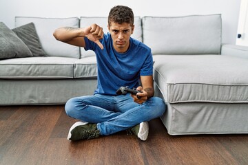 Young handsome hispanic man playing video game sitting by the sofa with angry face, negative sign showing dislike with thumbs down, rejection concept