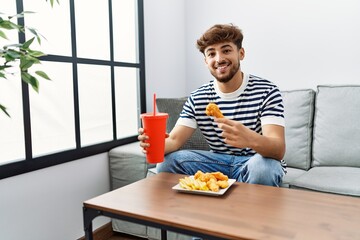 Young arab man smiling confident eating fried chicken drinking soda beverage at home