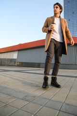 Low angle view of good looking man in brown coat holding coffee to go against buildings in city 