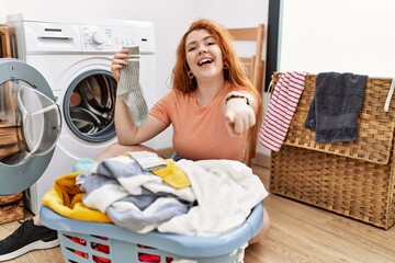 Young redhead woman putting dirty laundry into washing machine pointing to you and the camera with...