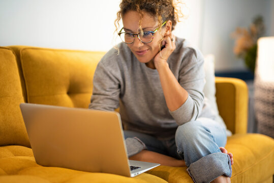 Happy young woman working on laptop sitting on sofa in living room at home. Businesswoman in casuals working from home. Caucasian woman in eyeglasses smiling while using laptop.