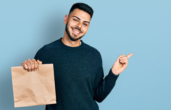 Young hispanic man with beard holding take away paper bag smiling happy pointing with hand and finger to the side