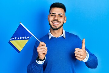 Young hispanic man with beard holding bosnia herzegovina flag smiling happy and positive, thumb up doing excellent and approval sign