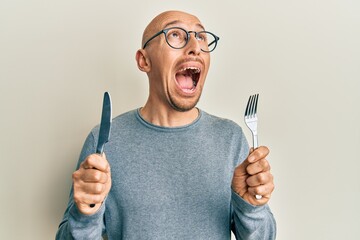 Bald man with beard holding fork and knife ready to eat angry and mad screaming frustrated and...