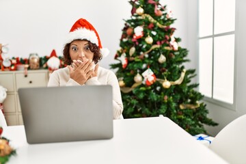 Middle age hispanic woman using laptop sitting by christmas tree shocked covering mouth with hands for mistake. secret concept.