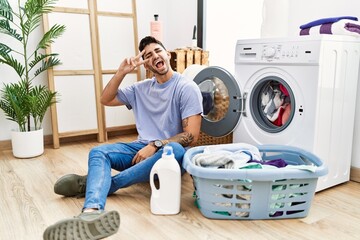 Young hispanic man putting dirty laundry into washing machine doing peace symbol with fingers over face, smiling cheerful showing victory