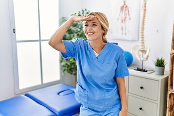 Fototapeta premium Beautiful blonde physiotherapist woman working at pain recovery clinic very happy and smiling looking far away with hand over head. searching concept.